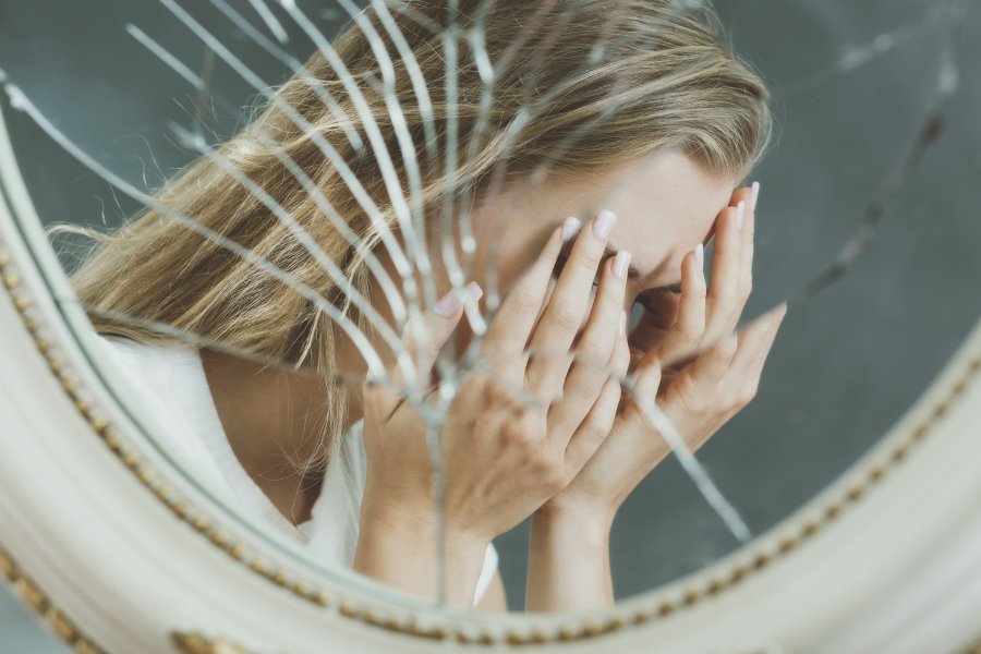 A woman's reflection in a shattered mirror, with her hands covering her face, visually encapsulates the 'Overcome Fear of Losing Someone' sentiment, suggesting the personal struggle and fragmented reality one faces with such fear.