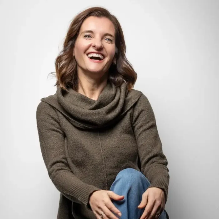 A woman with a joyful expression, wearing a brown cowl neck sweater and blue jeans, sitting with one leg crossed over the other against a white background.